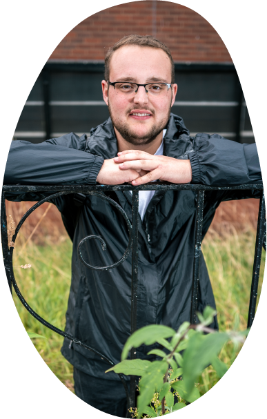 A young man smiling and looking hopeful, wearing glasses and an anorak, leaning on a gate