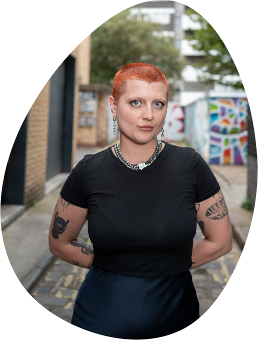 A woman with short orange hair, standing in a street with her arms behind her back, staring confidently forward