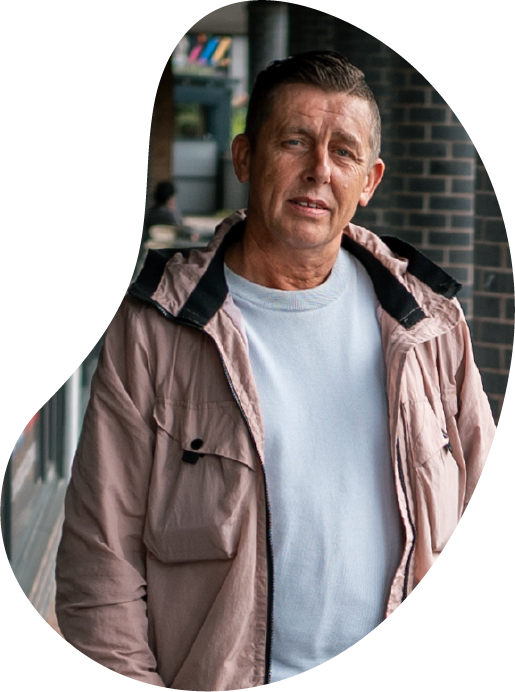 A photo of a man wearing a raincoat and light blue jumper, standing in an outdoor corridor