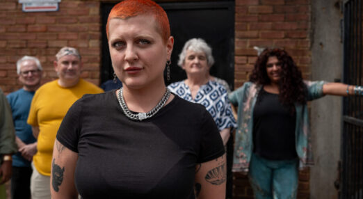 A photo of a woman staring straight ahead with her hands behind her back. Her friends are behind her