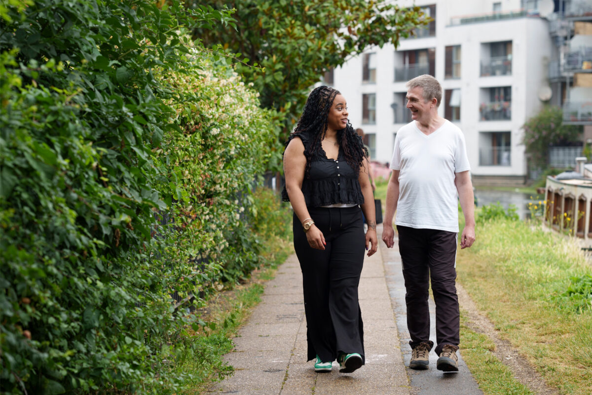 A woman and a man and walk along a canal path talking and smiling. There is grass on the path and a green, bush next to them.