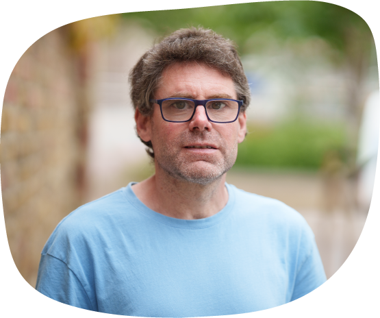 A photograph of Adam, someone who was supported by Waythrough's alcohol service in Haringey. He is stood in the street next to a wall. He has brown hair and is wearing a blue shirt
