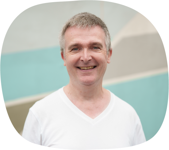 A photograph of Ian, who works in mental health support for Waythrough. He is wearing a white shirt, smiling, and standing in front of a wall with different pastel colours.
