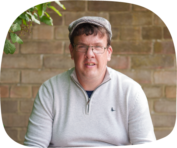 A photograph of Stephen, who was supported by Abbots Bank to live independently. He is sat on a bench, holding his phone, wearing glasses and a flat cap.
