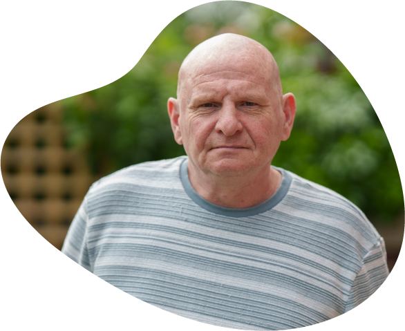A photograph of Ted, who was supported by Waythrough. He is stood in a leafy garden, wearing a striped shirt and looking straight at the camera.