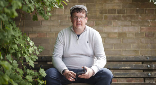 A photograph of Stephen, who was supported by Abbots Bank to live independently. He is sat on a bench, holding his phone, wearing glasses and a flat cap.