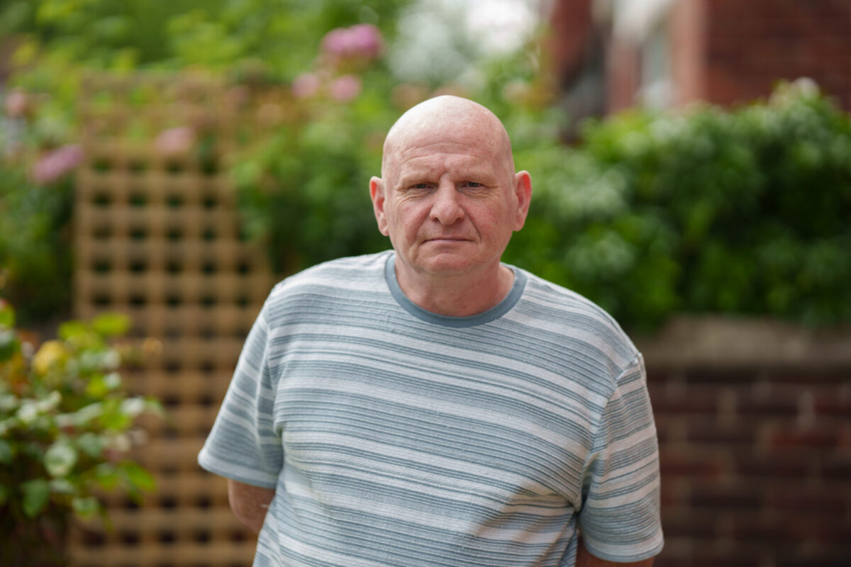 A photograph of Ted, who was supported by Waythrough. He is stood in a leafy garden, wearing a striped shirt and looking straight at the camera.