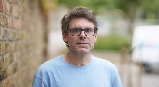 A photograph of Adam, someone who was supported by Waythrough's alcohol service in Haringey. He is stood in the street next to a wall. He has brown hair and is wearing a blue shirt