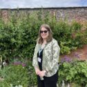 A photo of a woman in sunglasses stood in front of bushes, flowers and a brick wall.